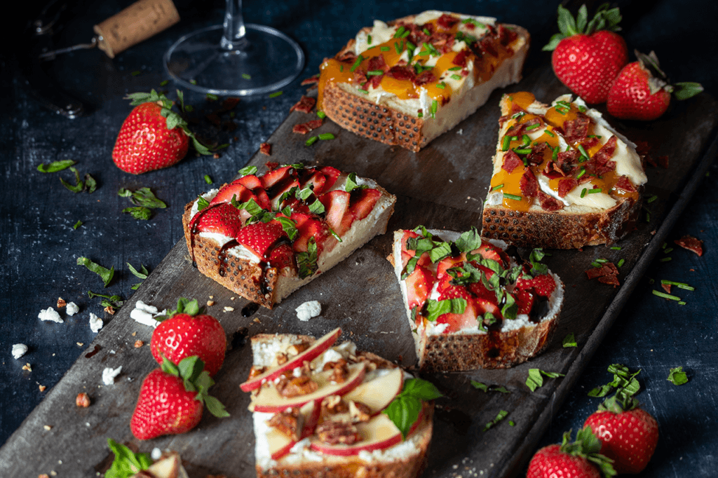 Front top view of pieces of different types of tomato and fruit bruschetta on a long wooden board.