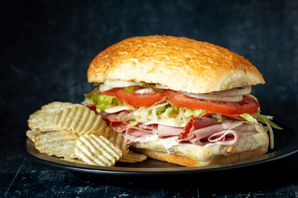 An italian sub tilted sideways on a black plate with a pile of potato chips next to it.